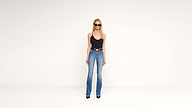 A woman wearing sunglasses, a black sleeveless top, blue high-waisted jeans, and black heels stands against a plain white background.