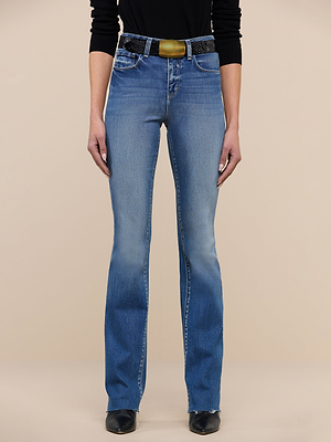 A person wears Ruth Straight-Leg Jeans in premium stretch denim, styled with a black top and a black belt with a large gold buckle, standing against a plain beige background. Only the body from shoulders down is shown.