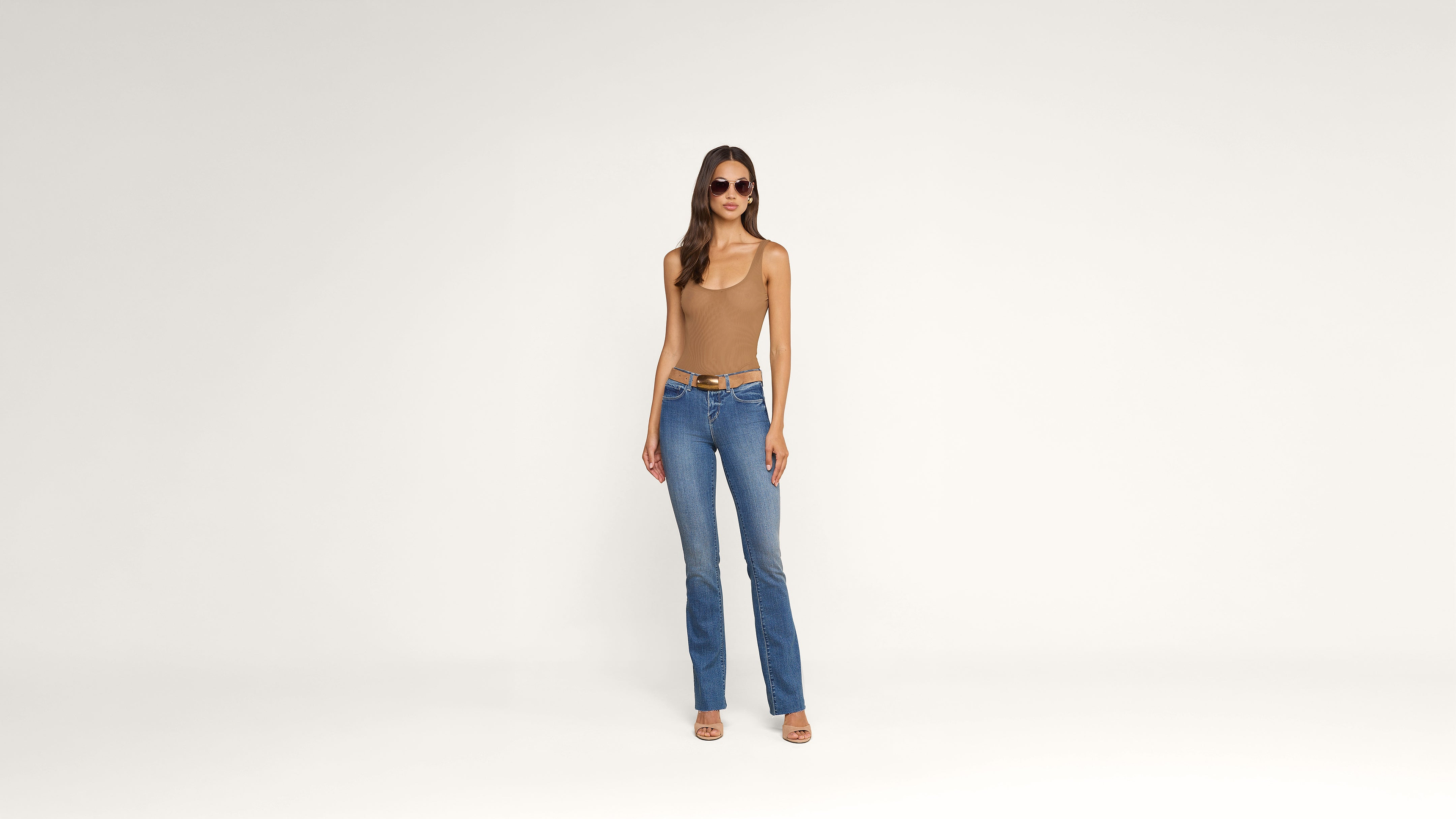 A woman wearing sunglasses, a tan sleeveless top, blue jeans, and sandals stands against a plain white background.