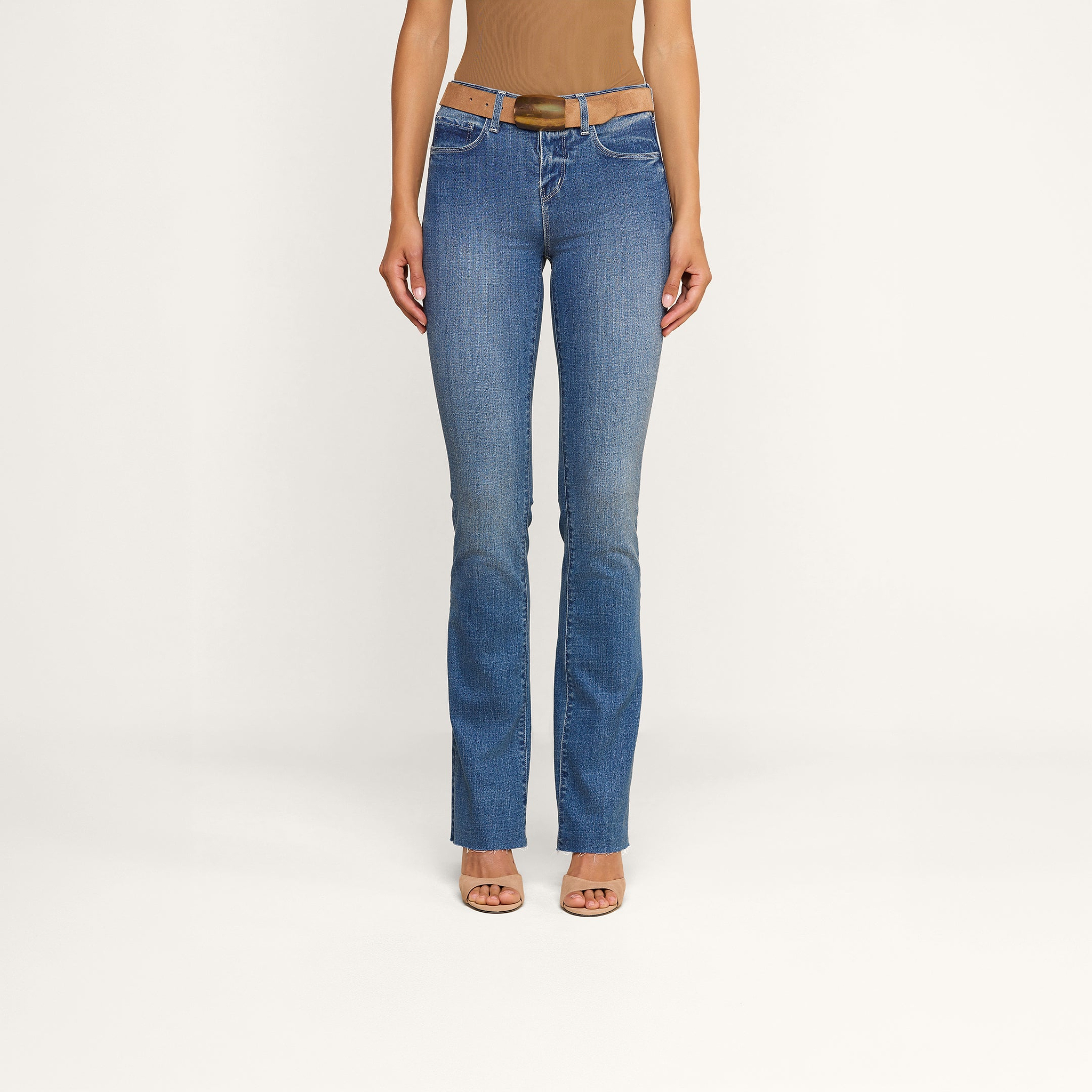 Una modella indossa i jeans Ruth Straight-Leg in classico indaco, con orlo grezzo e una leggera sfumatura sulla coscia, abbinati a una cintura marrone, un top senza maniche color cuoio e tacchi open-toe color carne, in piedi davanti a uno sfondo beige a tinta unita.