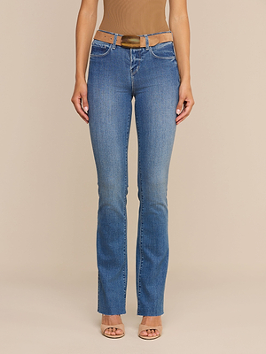 Una modella indossa i jeans Ruth Straight-Leg in classico indaco, con orlo grezzo e una leggera sfumatura sulla coscia, abbinati a una cintura marrone, un top senza maniche color cuoio e tacchi open-toe color carne, in piedi davanti a uno sfondo beige a tinta unita.