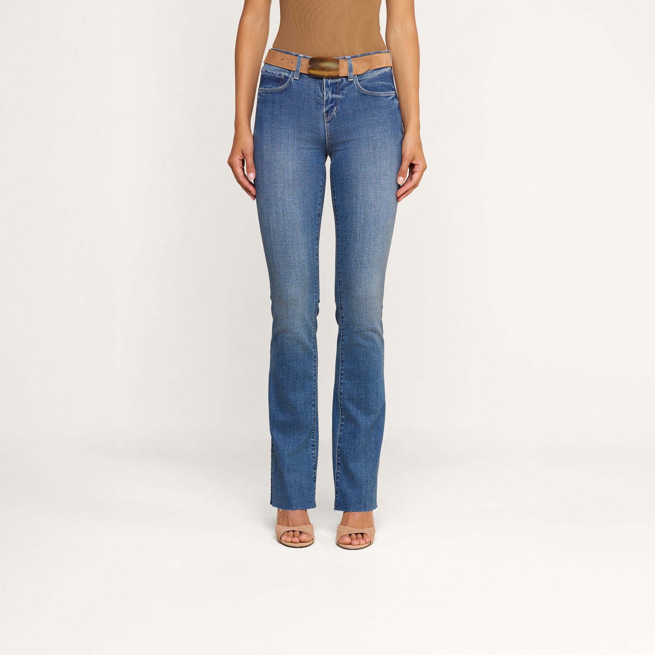 Una modella indossa i jeans Ruth Straight-Leg in classico indaco, con orlo grezzo e una leggera sfumatura sulla coscia, abbinati a una cintura marrone, un top senza maniche color cuoio e tacchi open-toe color carne, in piedi davanti a uno sfondo beige a tinta unita.