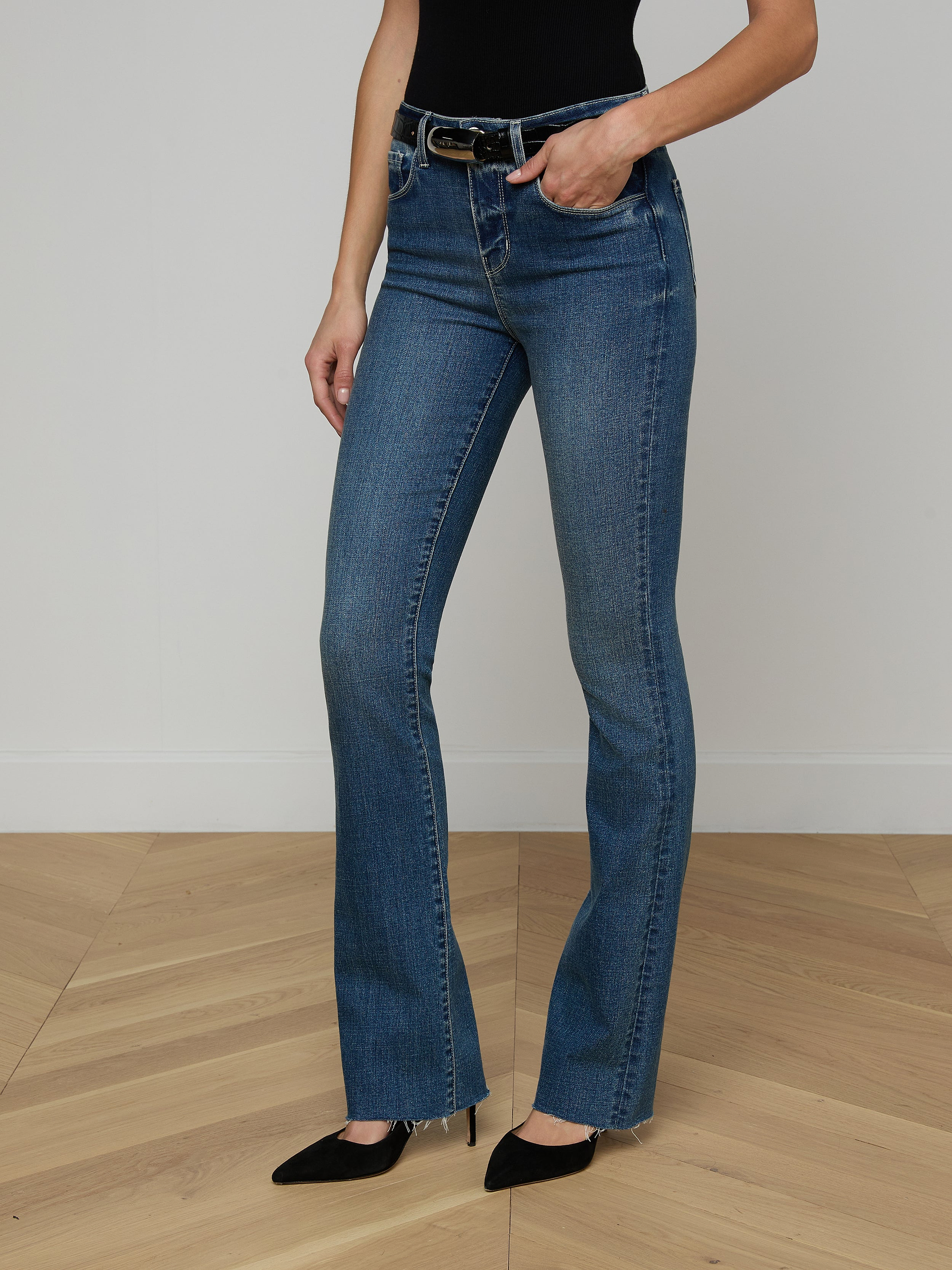 A person wearing the Ruth Straight-Leg Jean—a high-waisted indigo denim with a raw hem—paired with a brown sleeveless top and beige heeled sandals stands against a plain white background. Only the lower half of the body is visible.