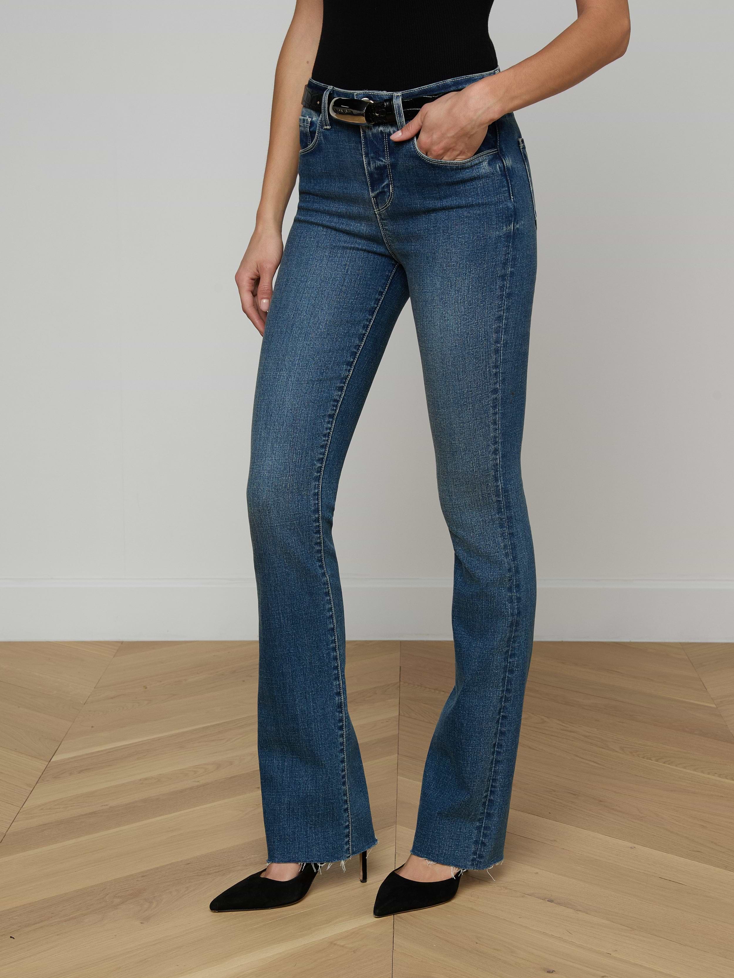A person wearing the Ruth Straight-Leg Jean—a high-waisted indigo denim with a raw hem—paired with a brown sleeveless top and beige heeled sandals stands against a plain white background. Only the lower half of the body is visible.
