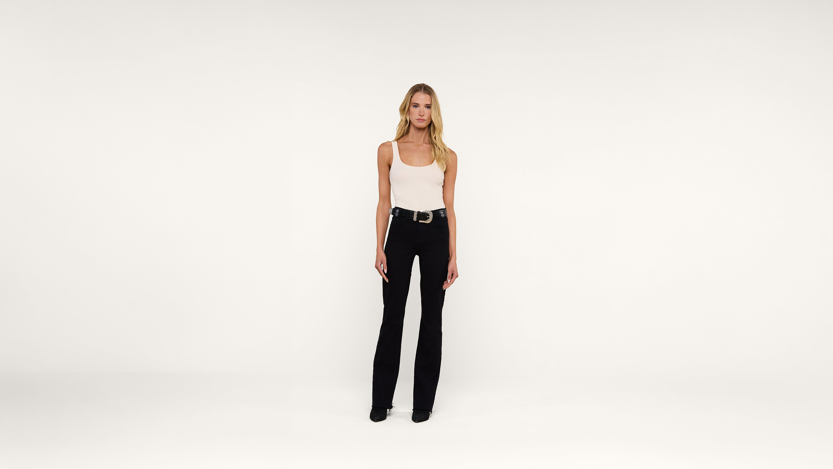 A woman with long blonde hair, wearing a white tank top, black pants, and black shoes stands facing forward in a plain, white studio setting.