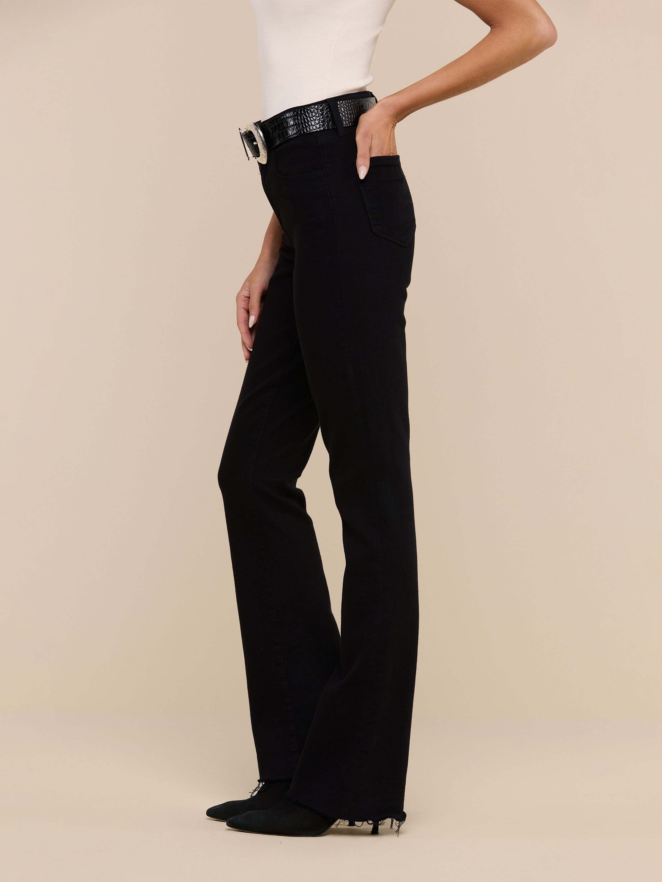 Woman wearing a sleeveless top, Ruth Straight-Leg Jean in black high-rise stretch denim, a black belt with a large buckle, and black shoes, standing against a plain white background.