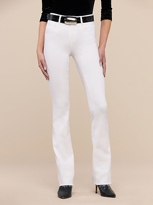 Wearing the Ruth Straight-Leg Jean in white premium stretch denim with a black belt, black top, and pointed-toe shoes, this person stands against a plain beige background.
