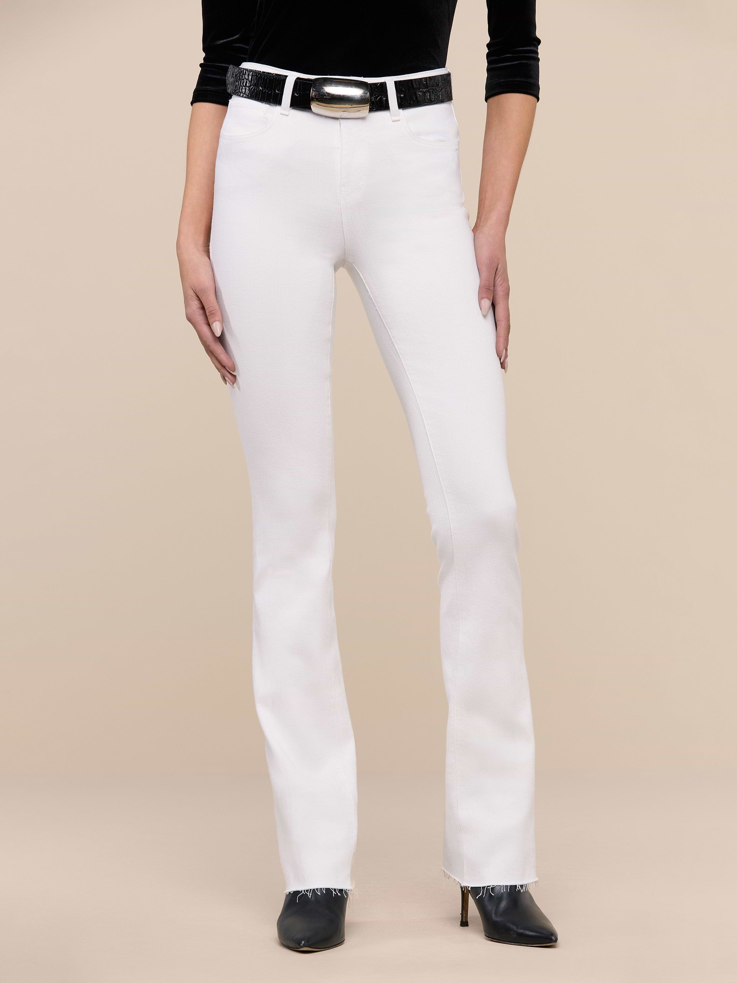 A person wearing the Ruth Straight-Leg Jean in white stretch denim, paired with a black top, a black belt with a silver buckle, and black pointed-toe heels stands against a plain white background.