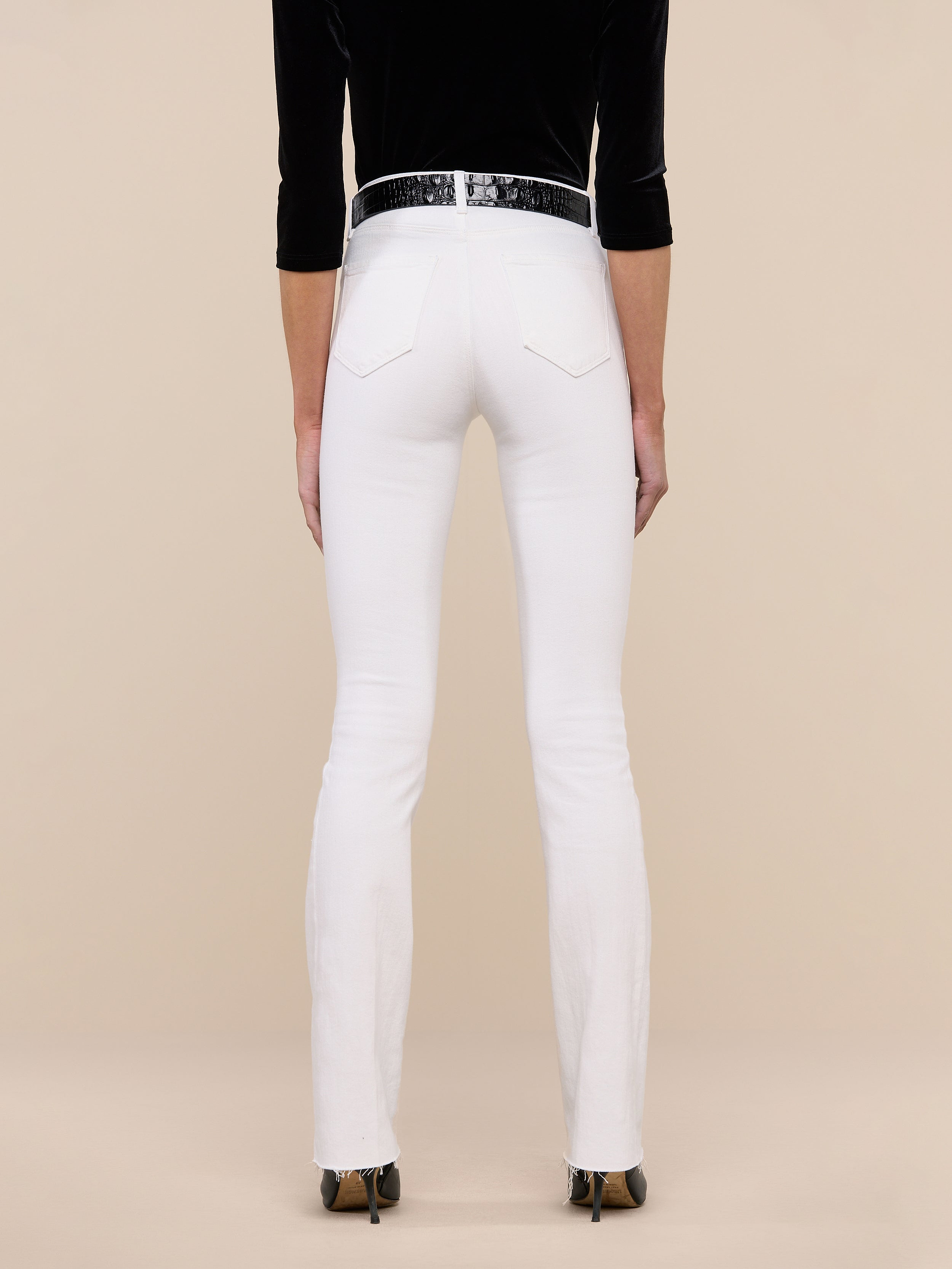 A person stands facing away against a plain white background, wearing the Ruth Straight-Leg Jean in white with a high-rise fit and slim straight leg, styled with a black belt, black long-sleeve top, and black-heeled shoes.