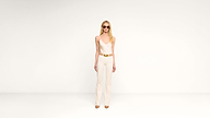 A woman wearing sunglasses, a white sleeveless top, white pants, a gold belt, and tan shoes stands against a plain white background.