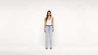 Woman standing against a plain white background, wearing a white tank top, light blue jeans, and tan shoes. She has long brown hair and faces the camera directly with a neutral expression.