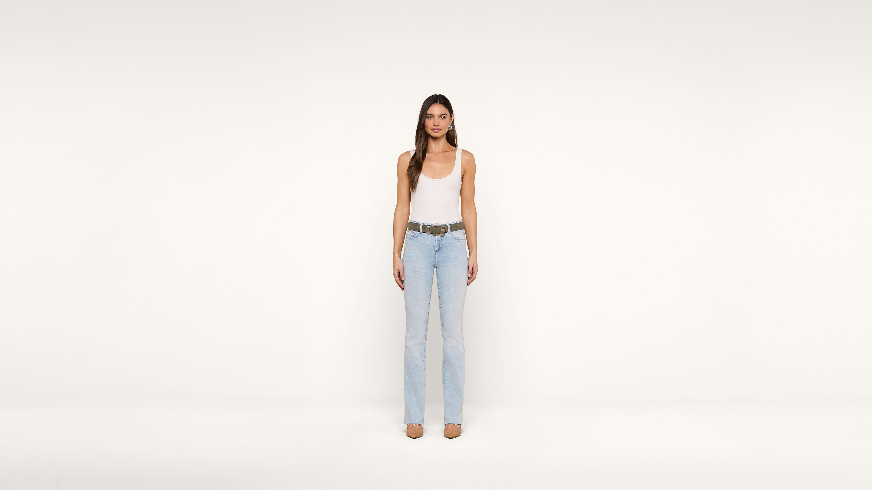 Woman standing against a plain white background, wearing a white tank top, light blue jeans, and tan shoes. She has long brown hair and faces the camera directly with a neutral expression.