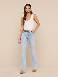 A woman stands against a plain white background wearing a white tank top, beige heels, and the Ruth Straight-Leg Jean in light blue classic stretch denim with a raw hem. She has long brown hair and a neutral expression, with one hand in her pocket.