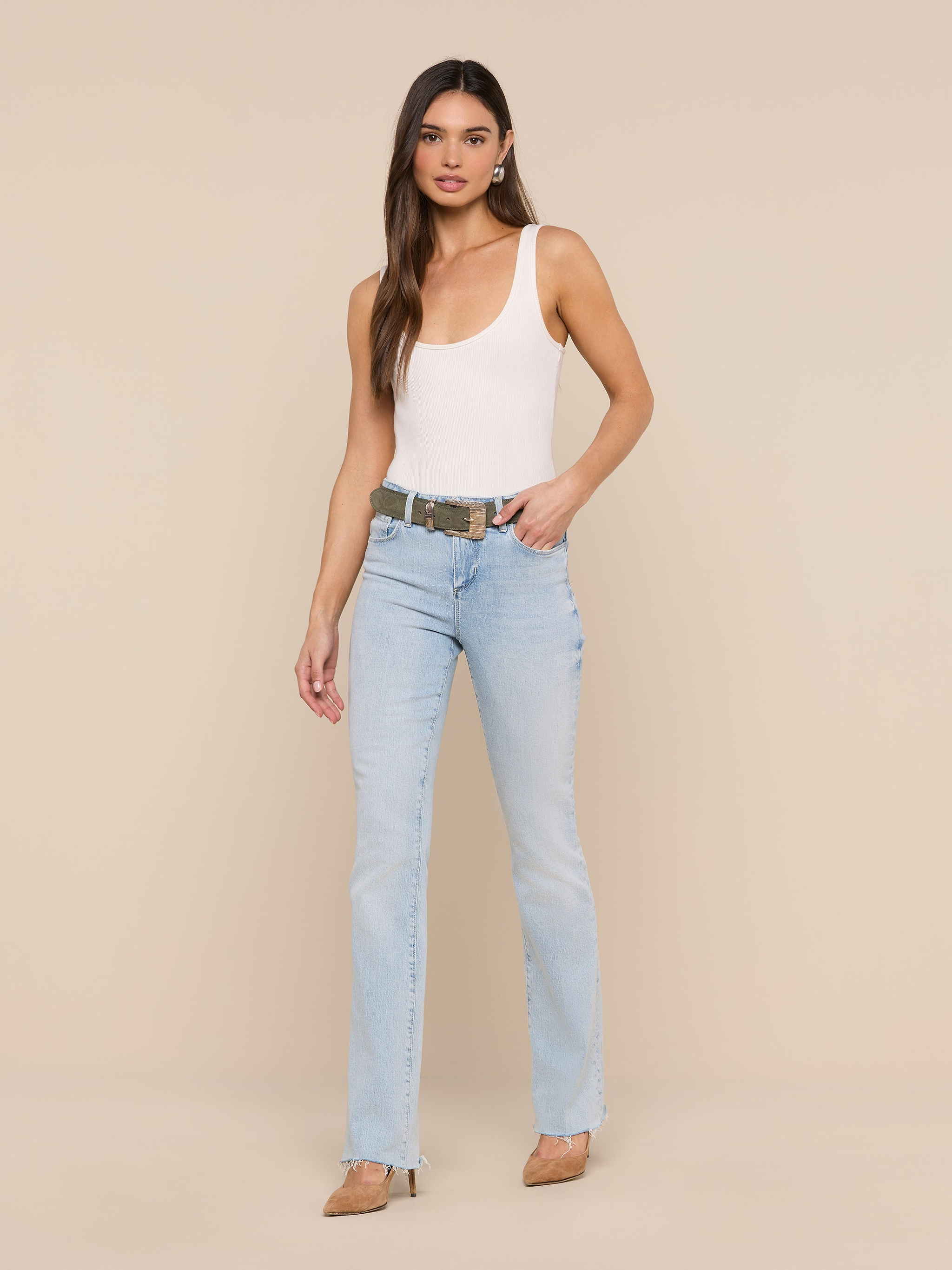 A woman stands against a plain white background wearing a white tank top, beige heels, and the Ruth Straight-Leg Jean in light blue classic stretch denim with a raw hem. She has long brown hair and a neutral expression, with one hand in her pocket.