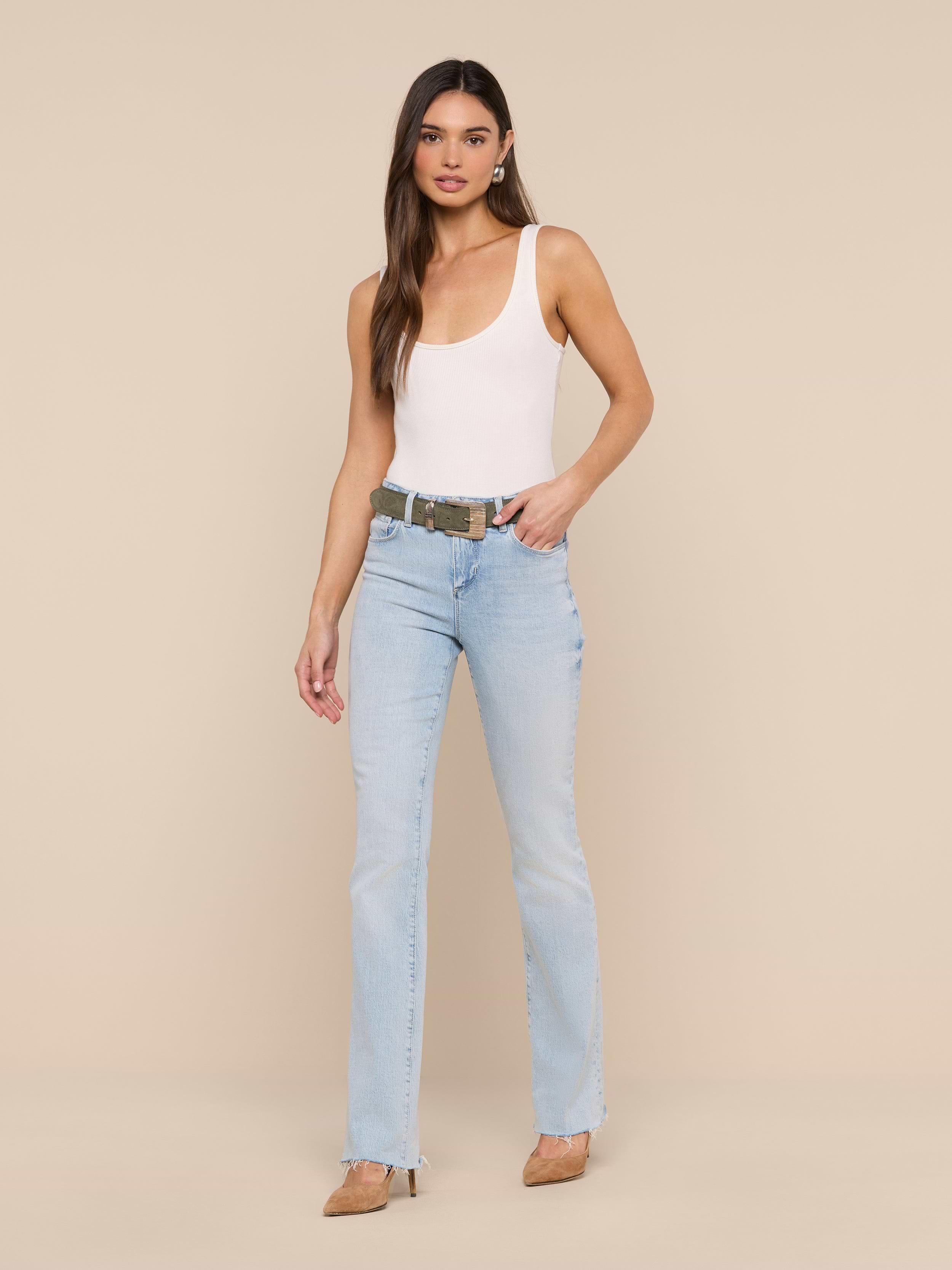 A woman stands against a plain white background wearing a white tank top, beige heels, and the Ruth Straight-Leg Jean in light blue classic stretch denim with a raw hem. She has long brown hair and a neutral expression, with one hand in her pocket.