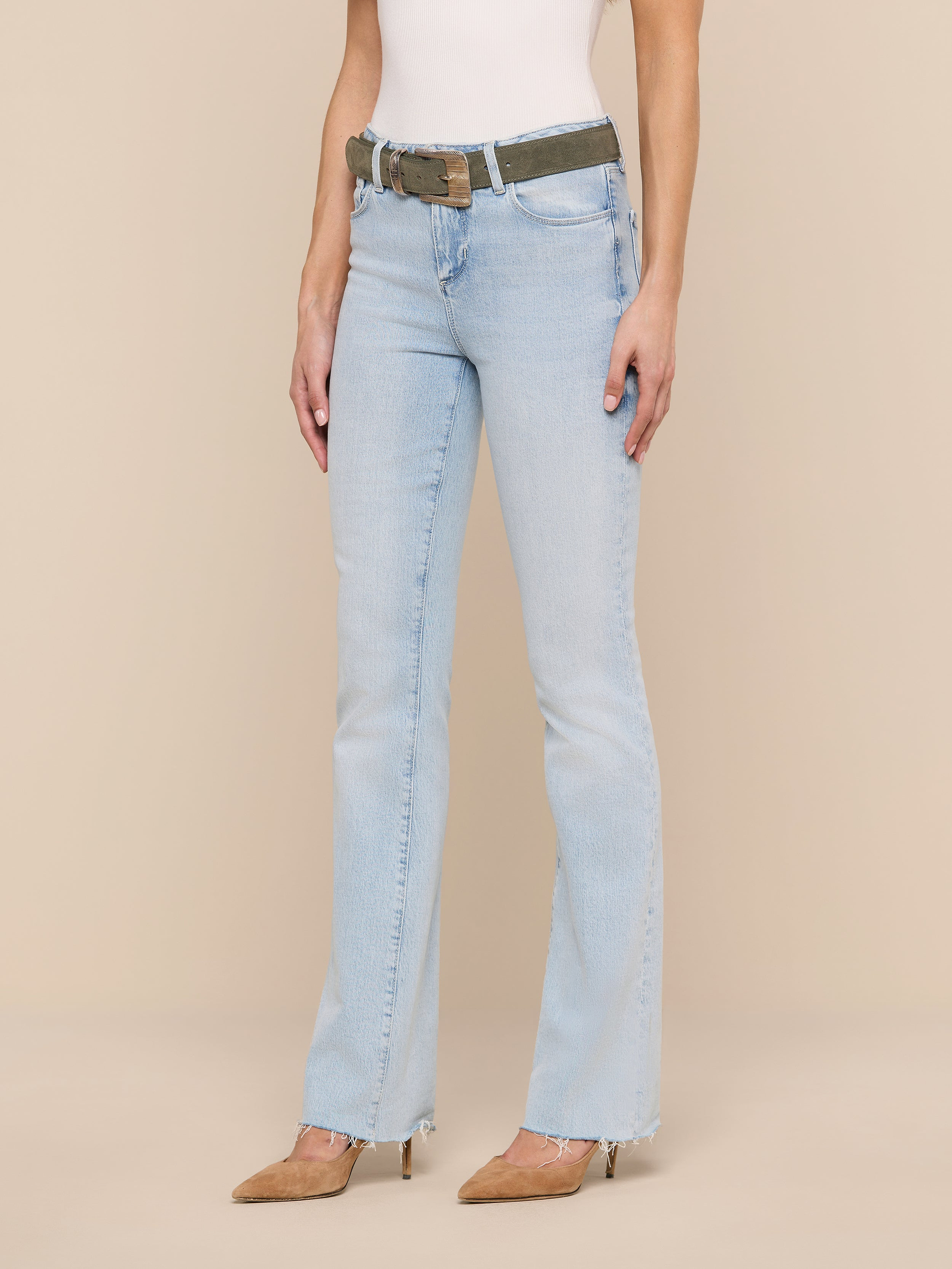 A woman wearing the Ruth Straight-Leg Jean in light blue with a raw hem, paired with a white sleeveless top, brown pointed-toe heels, and a camouflage belt, stands against a plain white background. Her head is not visible in the image.