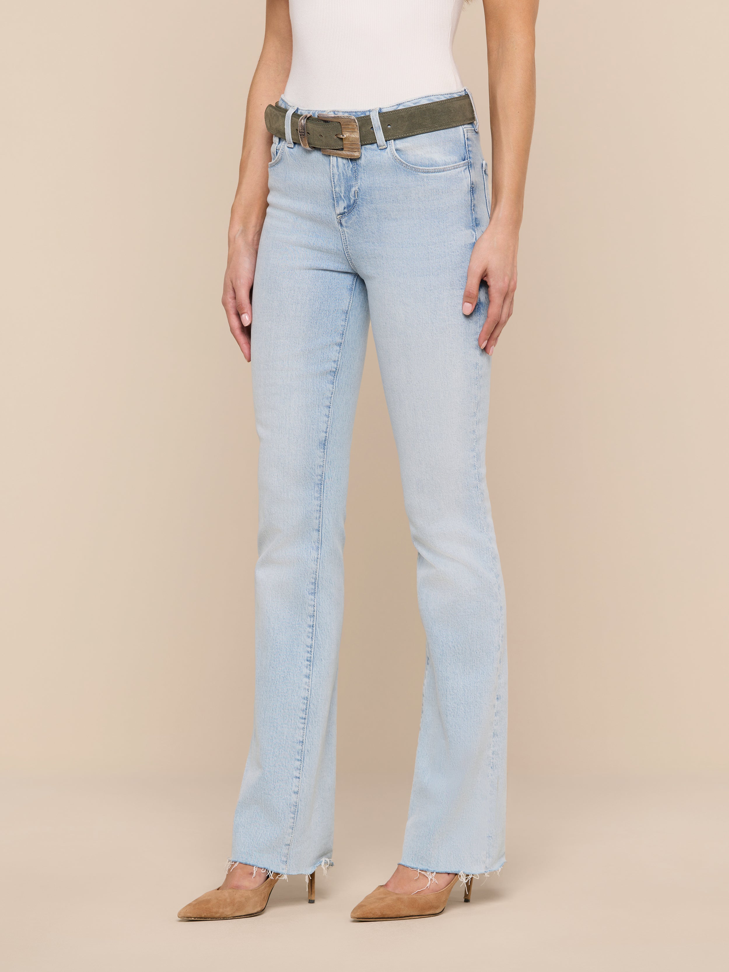 A woman wearing the Ruth Straight-Leg Jean in light blue with a raw hem, paired with a white sleeveless top, brown pointed-toe heels, and a camouflage belt, stands against a plain white background. Her head is not visible in the image.