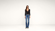 A woman wearing sunglasses, a black long-sleeve top with a deep V-neck, blue jeans, and black shoes stands against a plain white background.