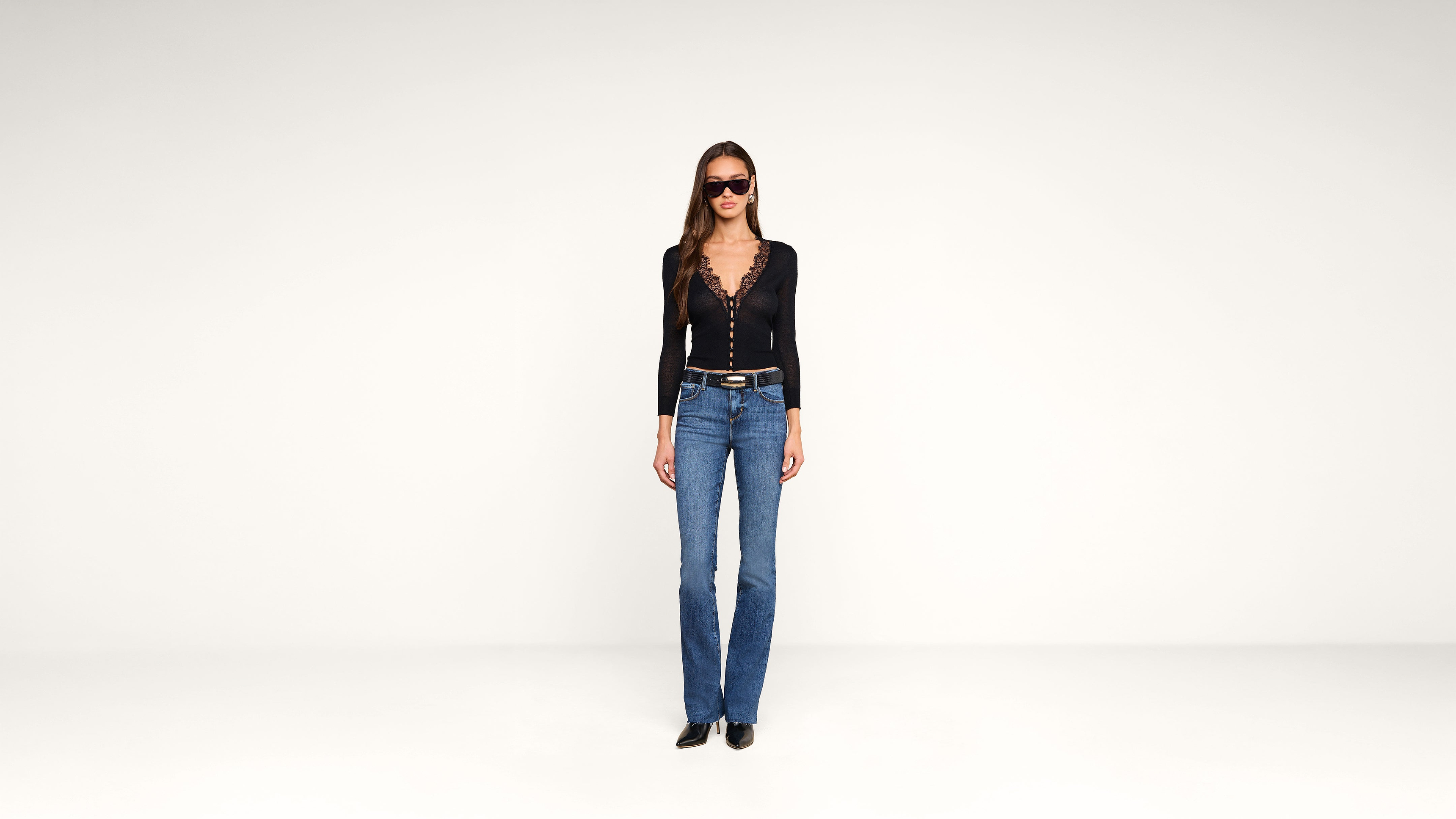 A woman wearing sunglasses, a black long-sleeve top with a deep V-neck, blue jeans, and black shoes stands against a plain white background.