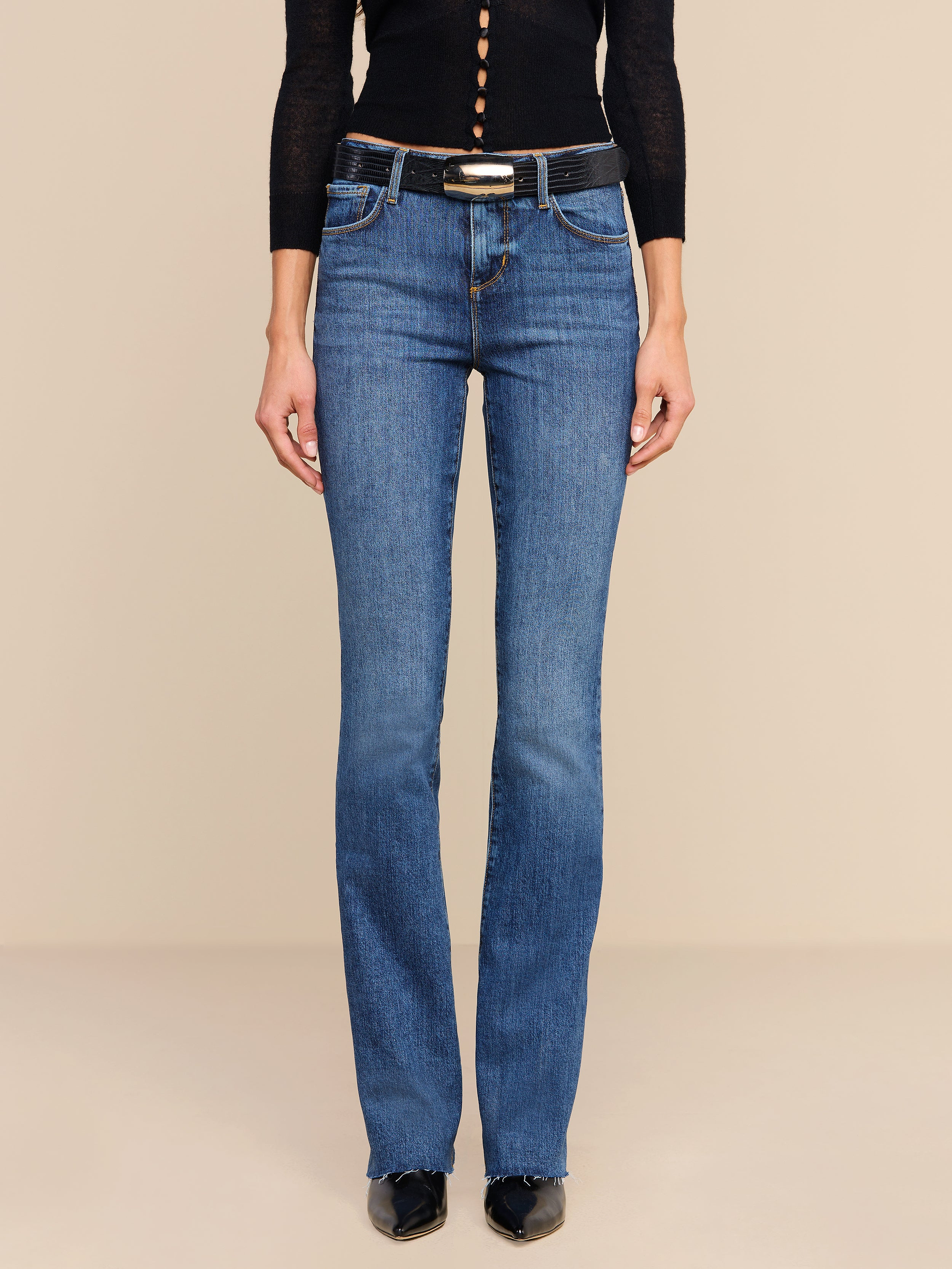 Wearing the Ruth Straight-Leg Jean in a medium indigo wash, styled with a black belt, black pointed shoes, and a black long-sleeve top, the model is shown from shoulders down against a plain white background.