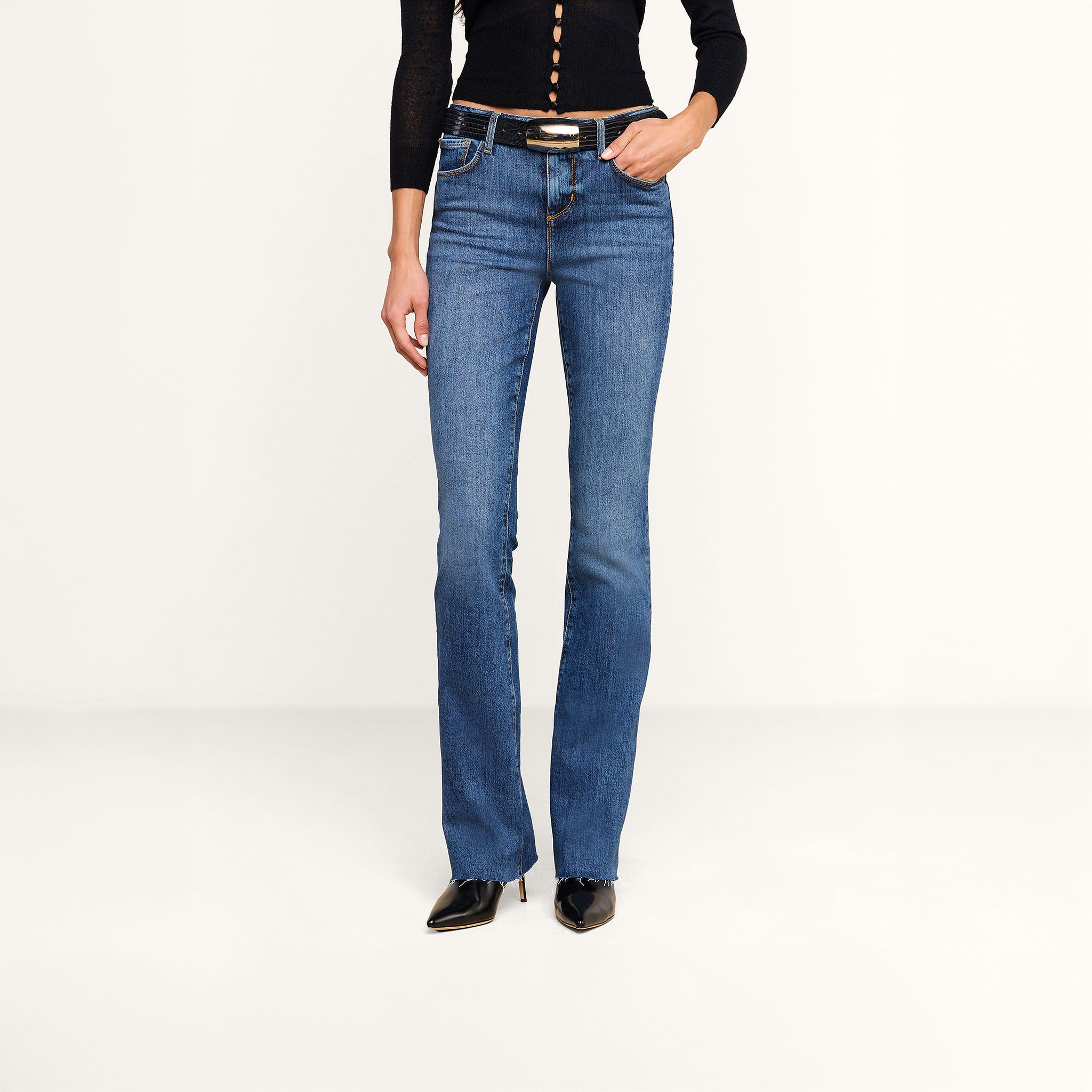 A person wearing a black long-sleeve top, the Ruth Straight-Leg Jean in medium indigo wash with a belt—crafted from stretch denim—and black pointed-toe shoes stands against a plain light background.