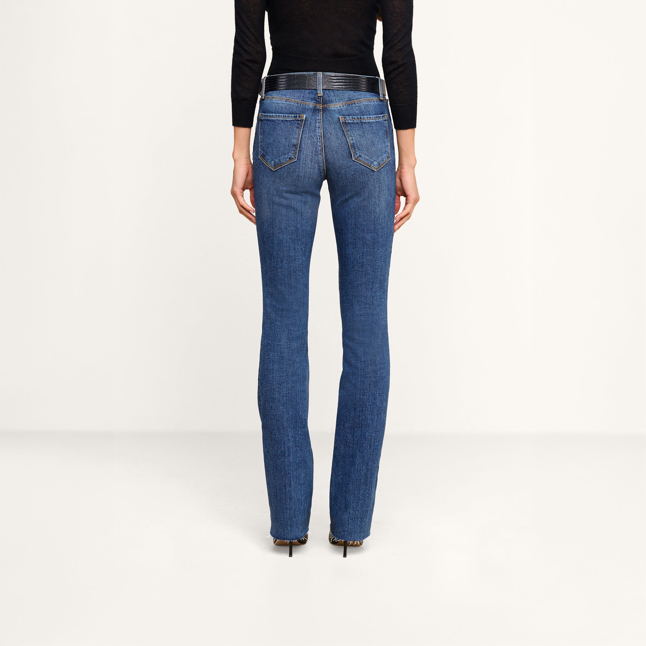 A person is shown from the back against a plain background, wearing a black long-sleeve top, black heels, and the Ruth Straight-Leg Jean in medium indigo wash with a fitted silhouette made from stretch denim.