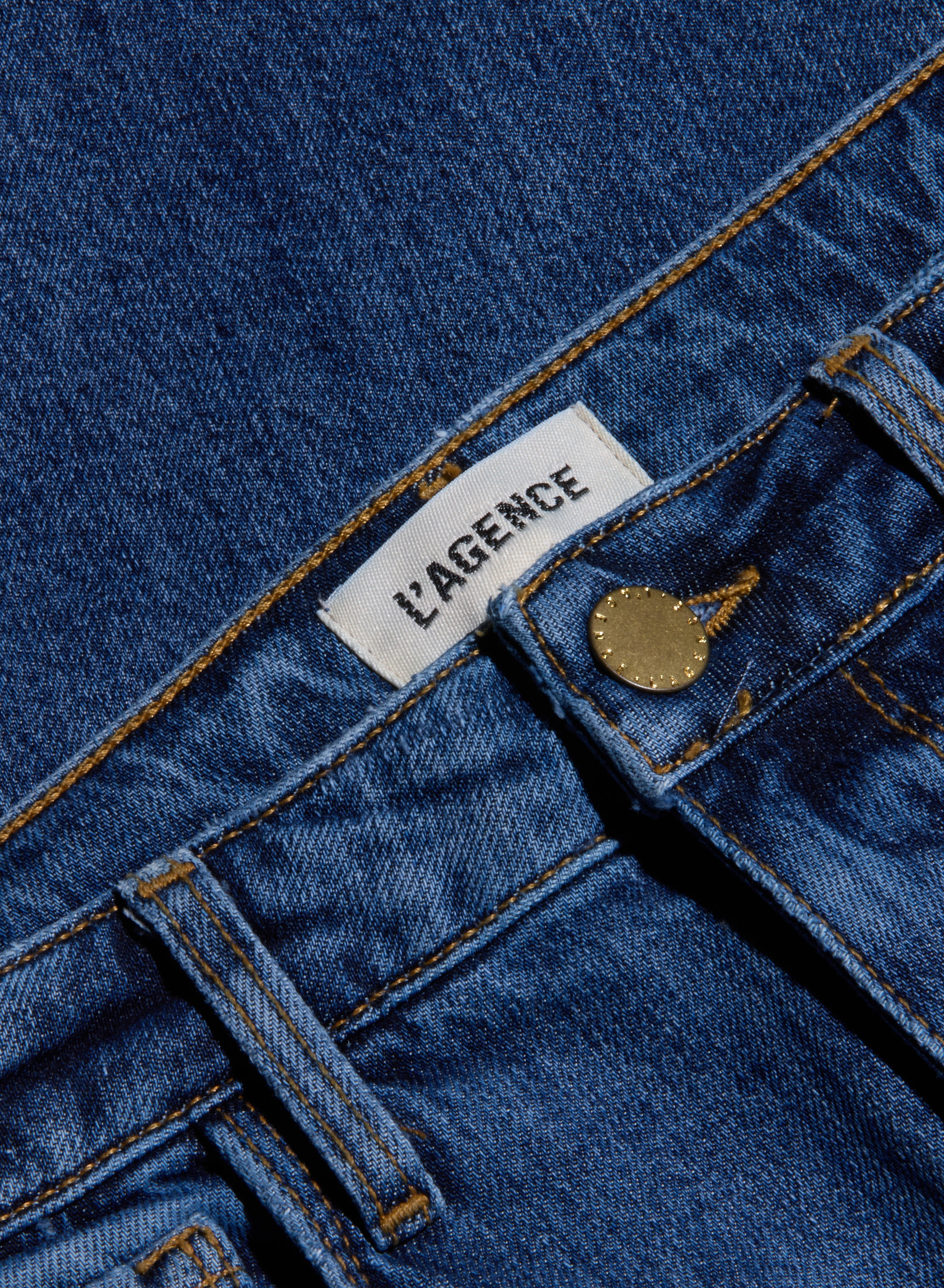 Close-up of blue stretch denim Ruth Straight-Leg Jean featuring a white LAGENCE label, a front belt loop, and a brass button. This fitted style comes in a medium indigo wash.