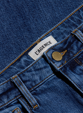 Close-up of the Ruth Straight-Leg Jean in a medium indigo wash, featuring blue stretch denim, a visible white L’AGENCE label, and a gold button at the waistband. This fitted jean offers both comfort and style with its straight-leg silhouette.