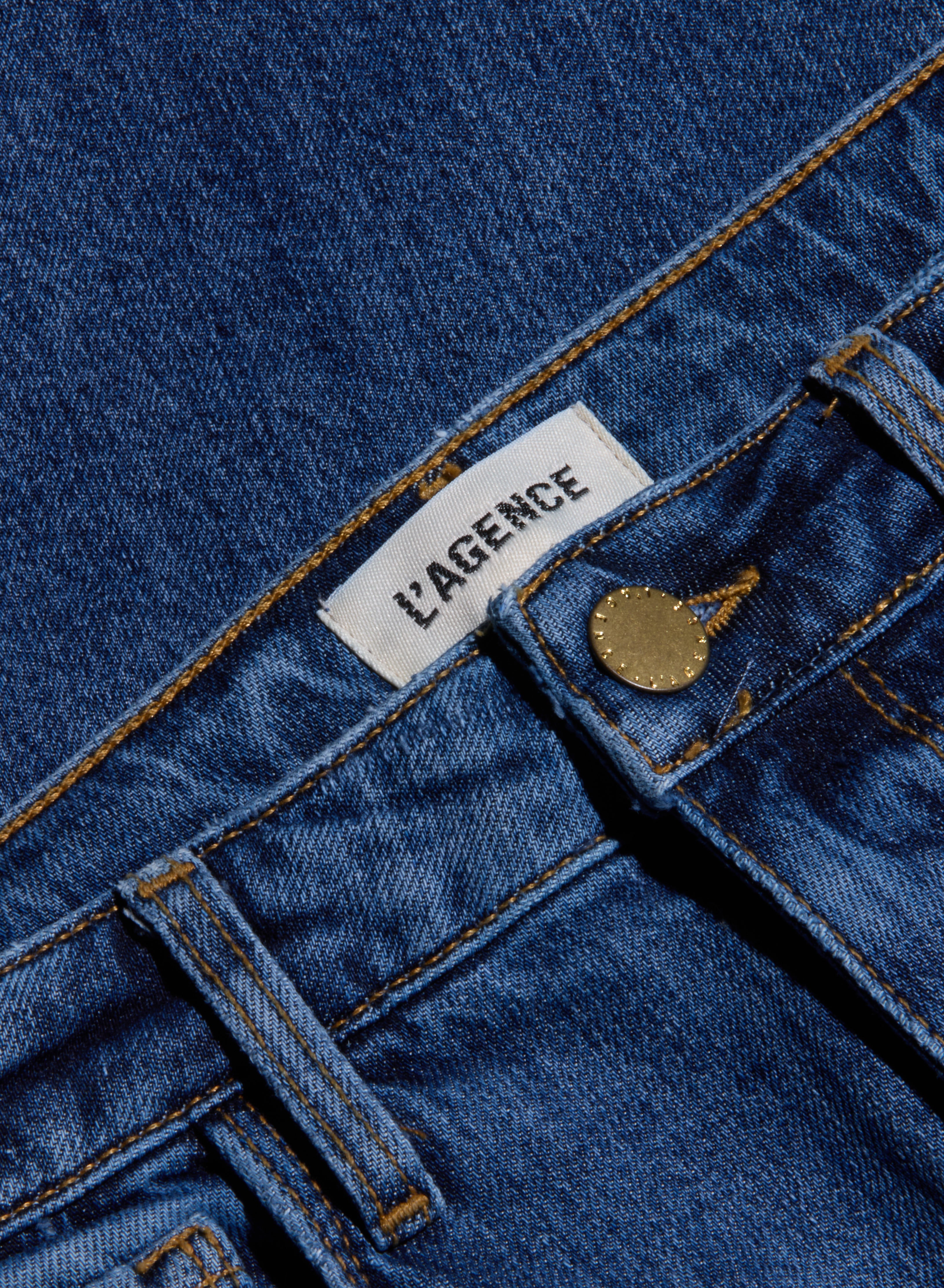 Close-up of blue stretch denim Ruth Straight-Leg Jean featuring a white LAGENCE label, a front belt loop, and a brass button. This fitted style comes in a medium indigo wash.