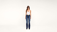 A woman wearing sunglasses, a white tank top, blue jeans, and black shoes stands against a plain white background.