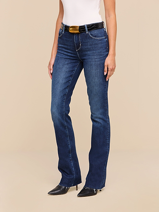 A woman wearing the Ruth Straight-Leg Jean in an indigo wash, paired with a white sleeveless top, a black belt featuring a gold buckle, and black pointed-toe shoes, stands against a beige background. Her face is not visible.