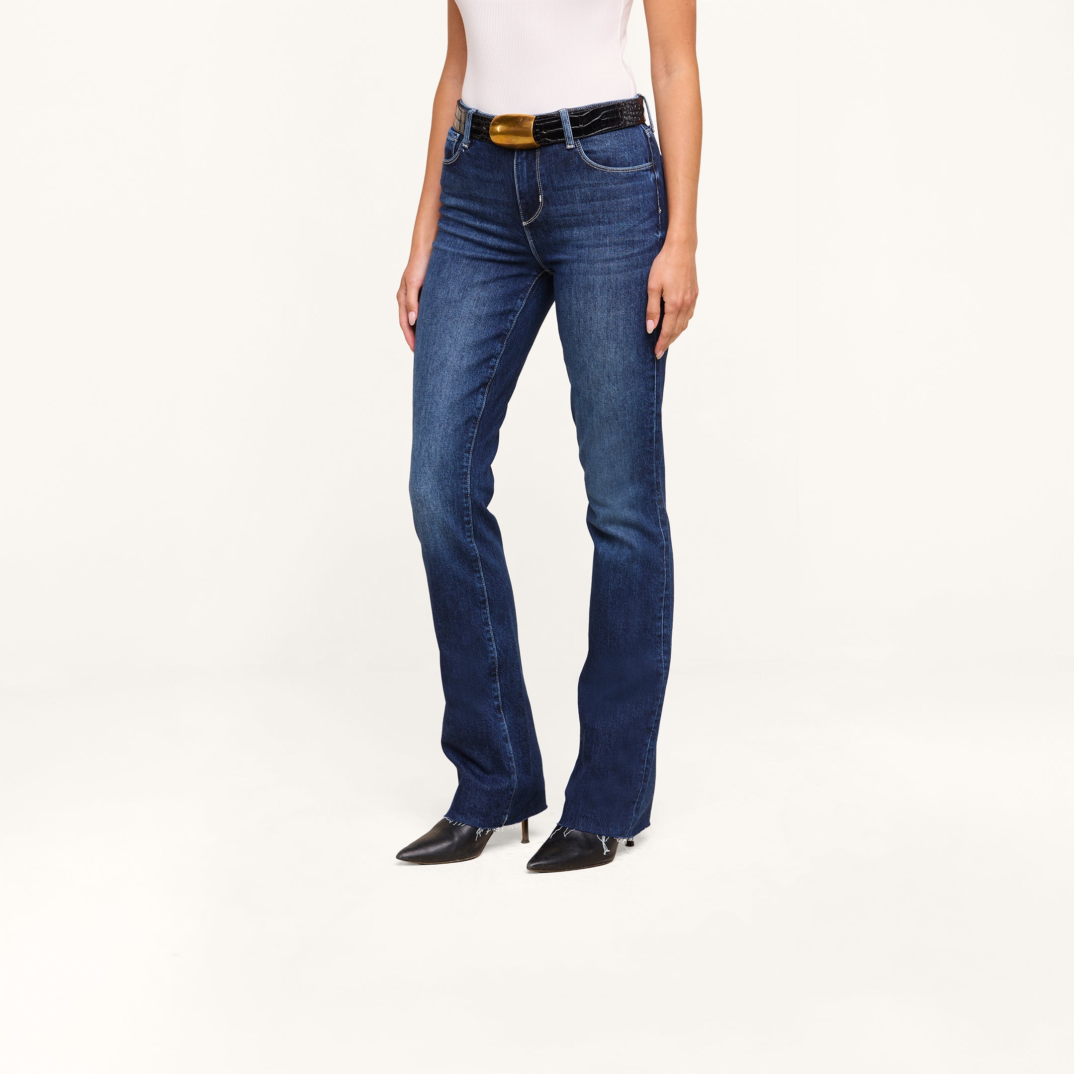 A person wearing Ruth Straight-Leg Jeans in dark blue, paired with a black belt featuring a gold buckle, black pointed shoes, and a white top stands against a plain white background.