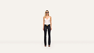 A woman stands against a plain white background, wearing sunglasses, a white sleeveless top, black pants, and trendy black heeled sandals.