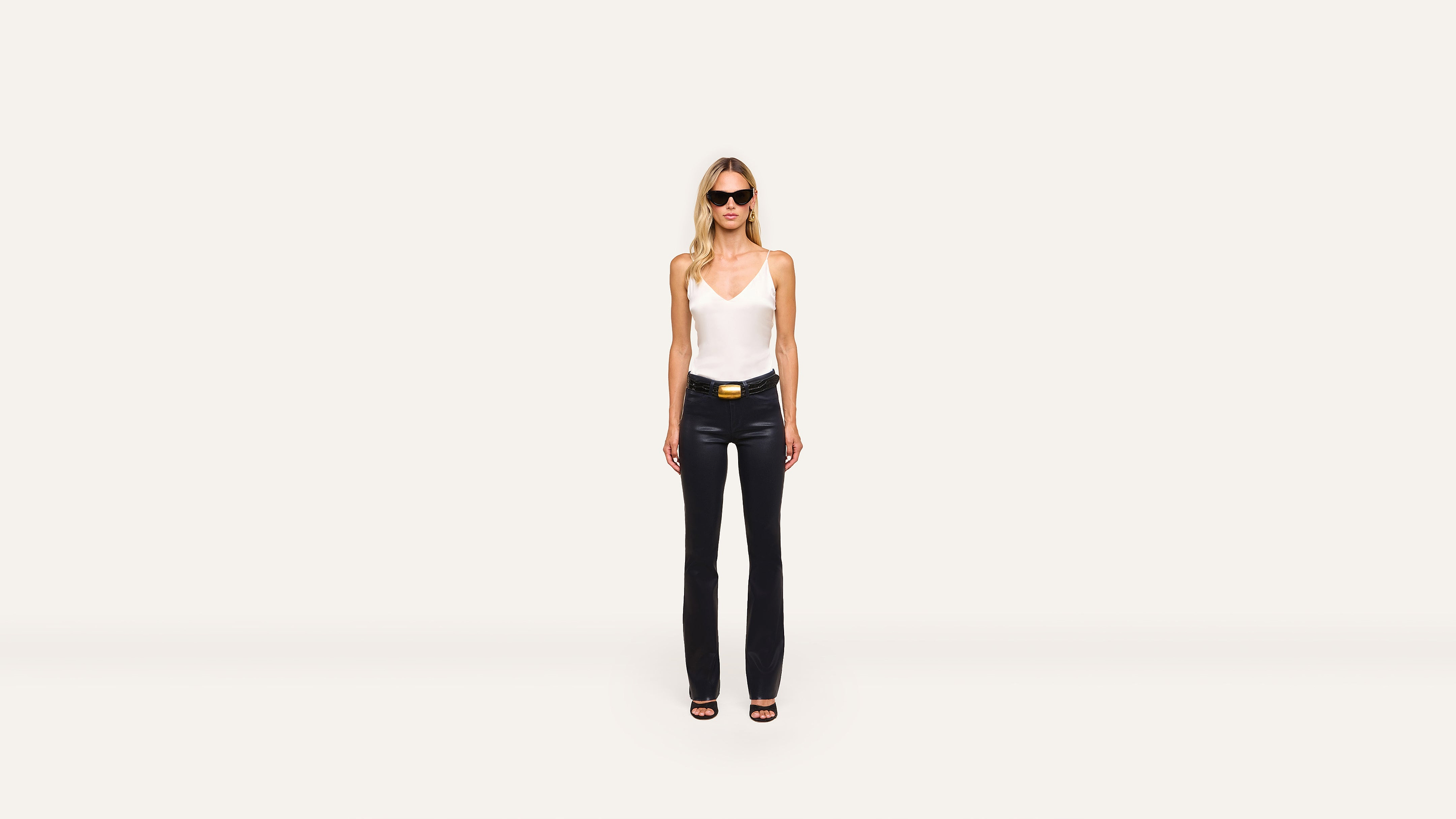 A woman stands against a plain white background, wearing sunglasses, a white sleeveless top, black pants, and trendy black heeled sandals.