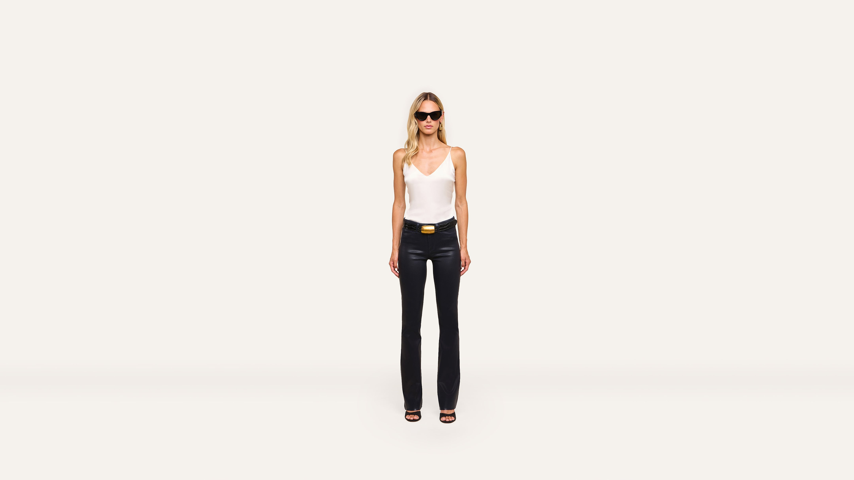 A woman stands against a plain white background, wearing sunglasses, a white sleeveless top, black pants, and trendy black heeled sandals.