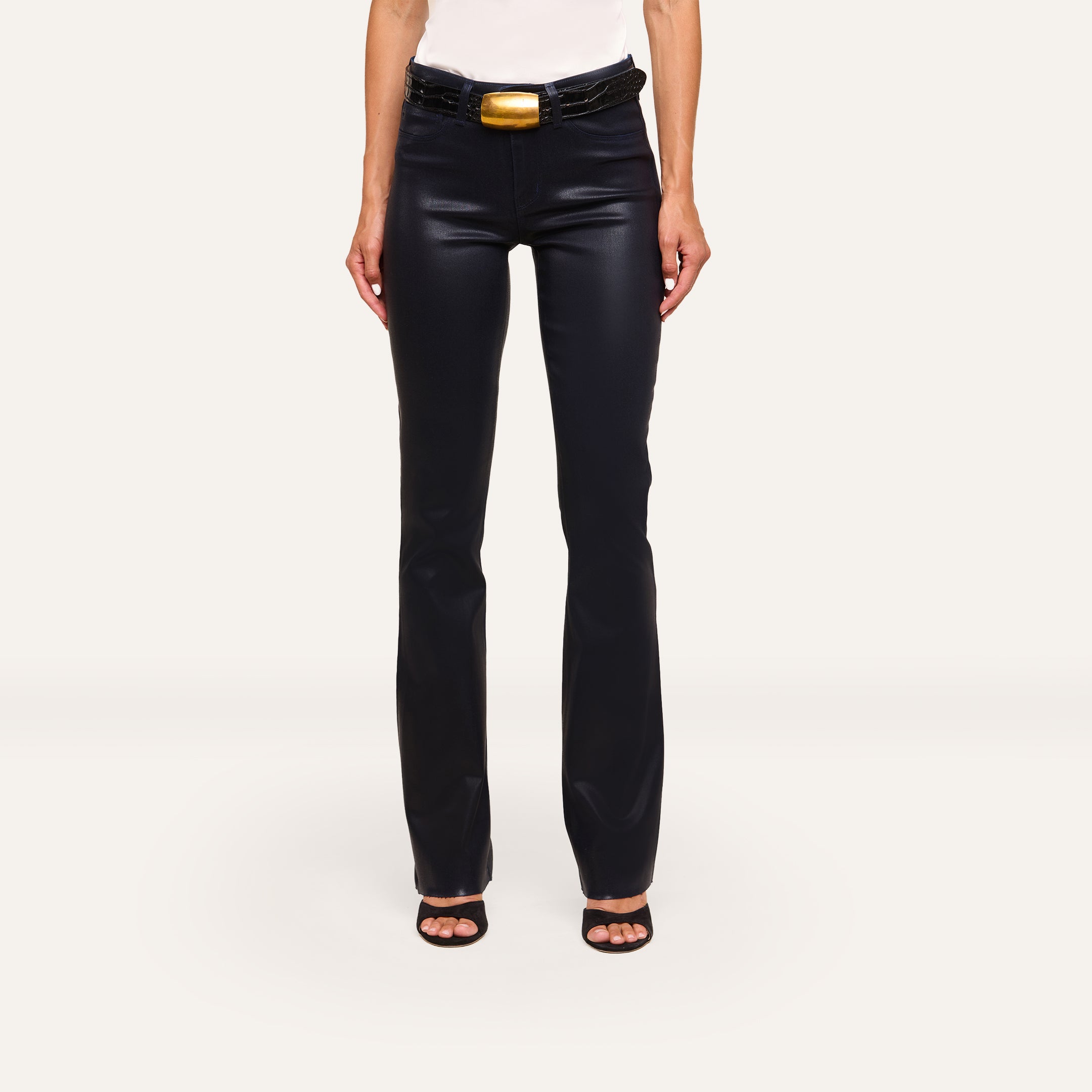A person wears Ruth Coated Straight-Leg Jean in black, paired with a black belt featuring a large gold buckle, black heeled sandals, and a white top, standing against a plain white background.