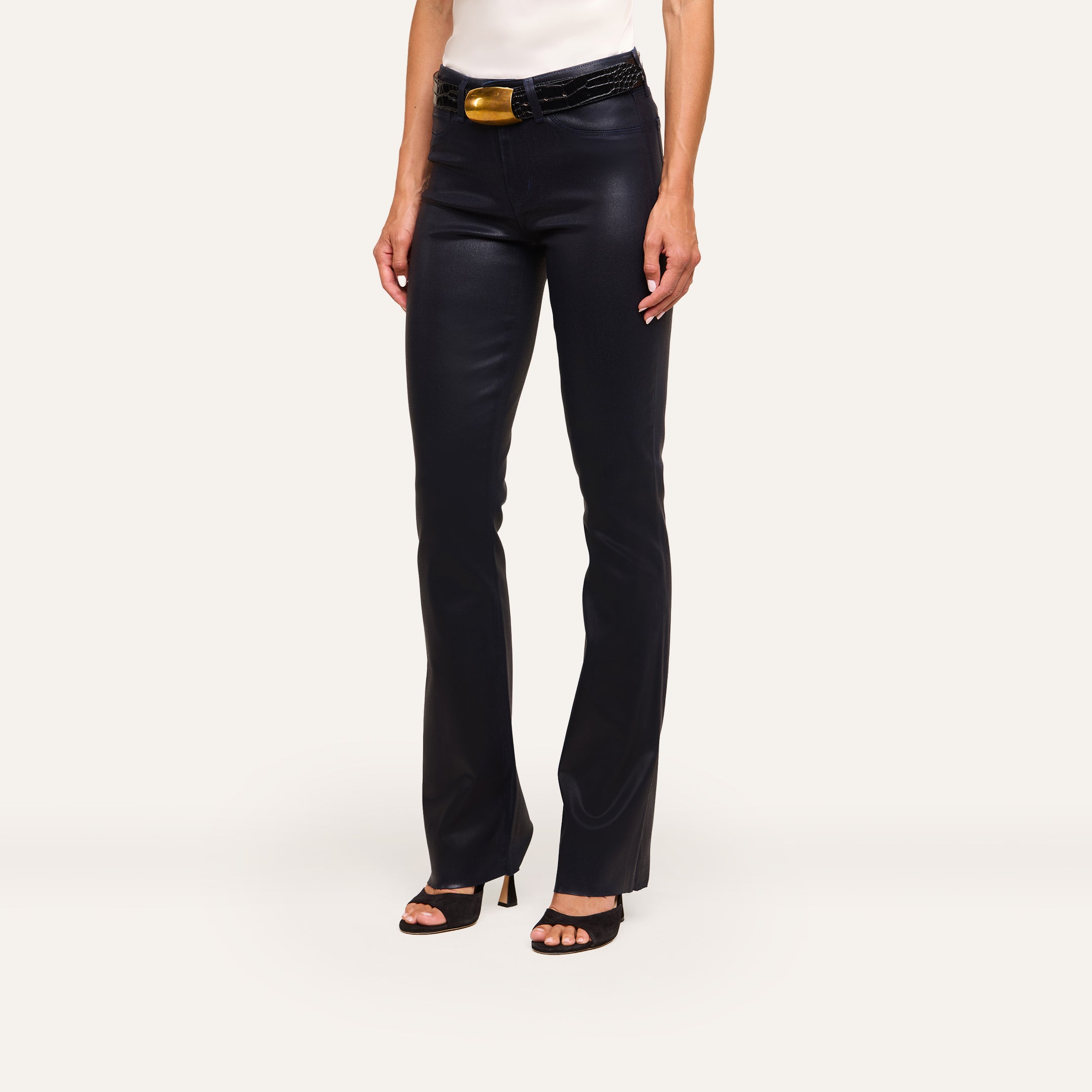 A person stands against a plain light background wearing Ruth Coated Straight-Leg Jean in black, a white sleeveless top, a wide black belt with a large gold buckle, and black heeled sandals.