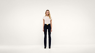 A woman with long blonde hair stands facing forward against a plain white background, wearing a white top, black pants, a belt, and black shoes.