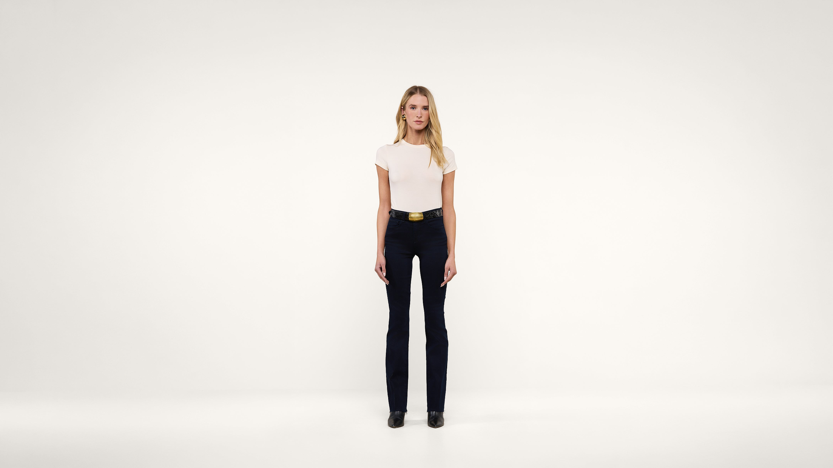 A woman with long blonde hair stands facing forward against a plain white background, wearing a white top, black pants, a belt, and black shoes.