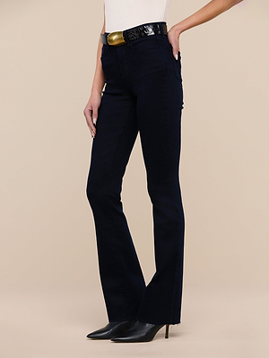 A person wearing Ruth Straight-Leg Jeans in ultra dark indigo wash, a black belt with a gold buckle, a white top, and black pointed-toe heeled boots stands against a plain beige background.