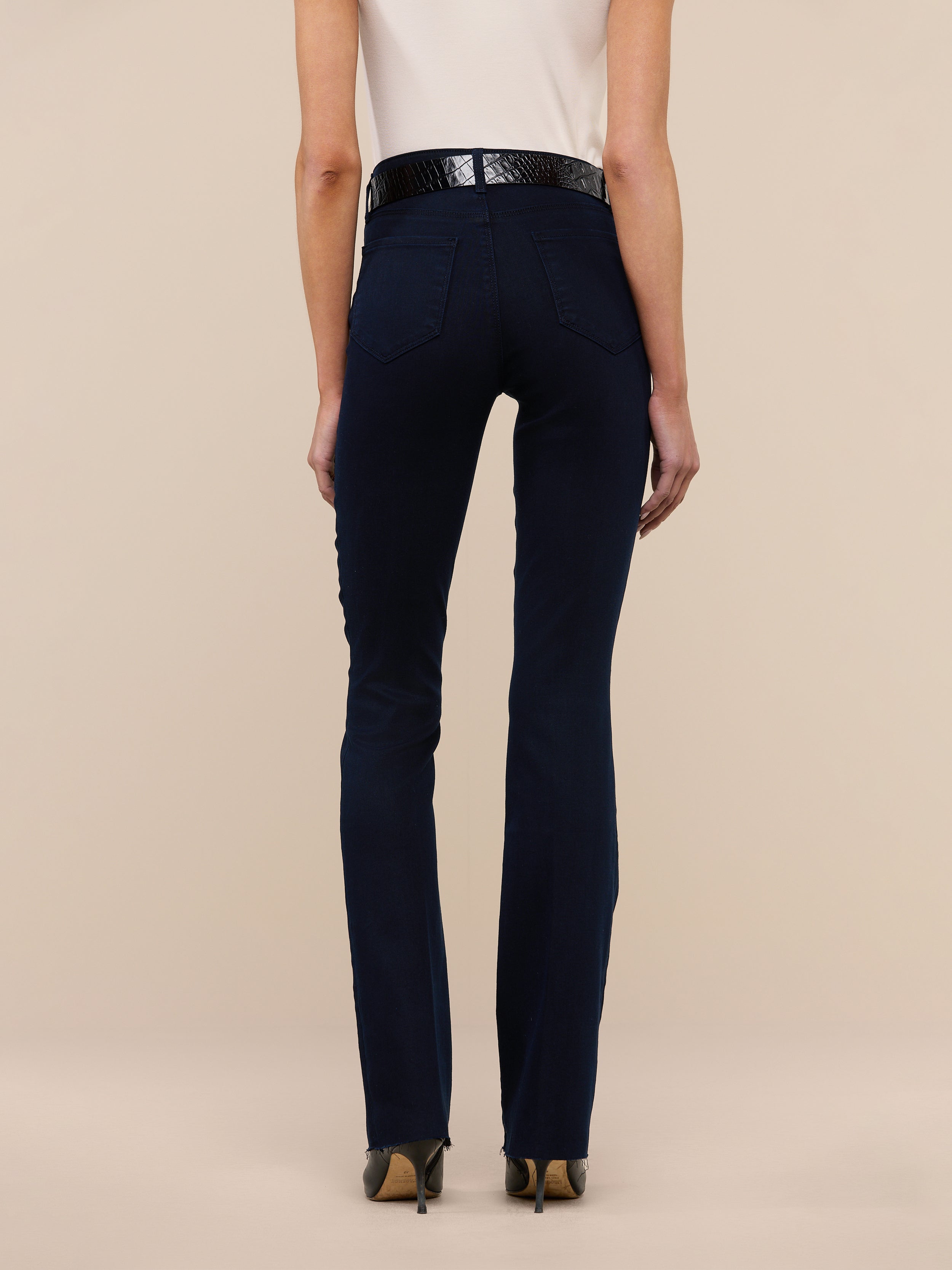 A person stands facing away, wearing a white sleeveless top, the Ruth Straight-Leg Jean in an ultra dark indigo wash, a black belt, and tan heels against a plain white background.
