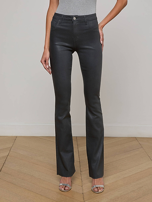 A person wears L'AGENCE Ruth Coated Straight-Leg Jeans, a gray top, and silver open-toe heels, standing on a light wood floor against an off-white wall. The person's head is not visible.