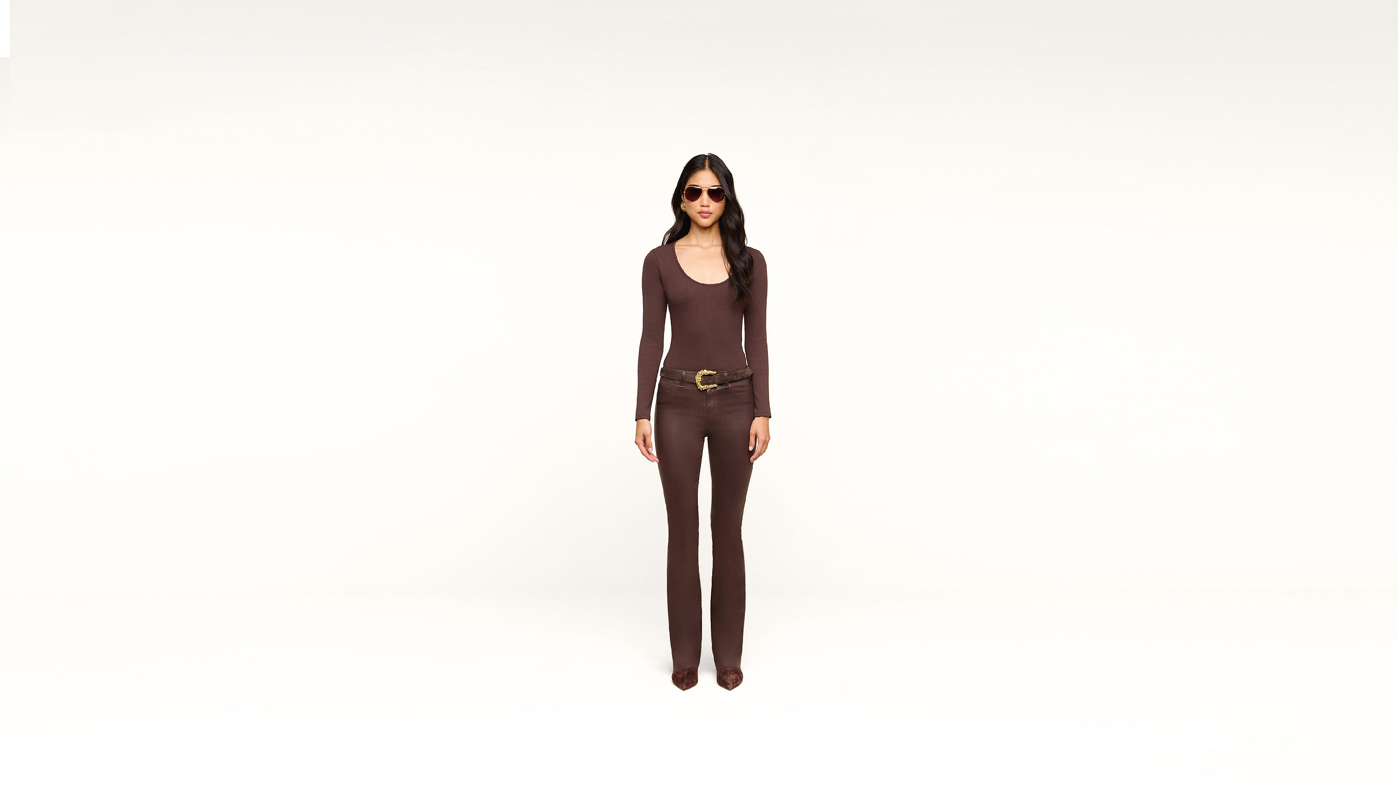 A woman wearing sunglasses, a fitted brown long-sleeve top, matching brown pants, and a large belt stands against a plain white background.