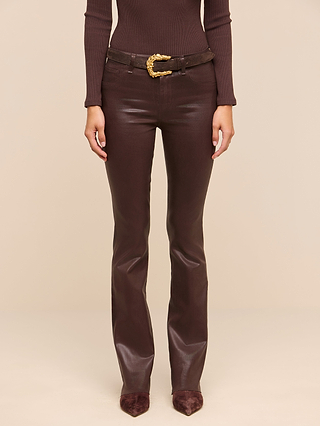 A person wearing the Ruth Coated Straight-Leg Jean—dark brown, shiny leather pants with a raw hem—styled with a matching long-sleeve top, pointed shoes, and a brown belt featuring a decorative gold buckle stands against a beige background.
