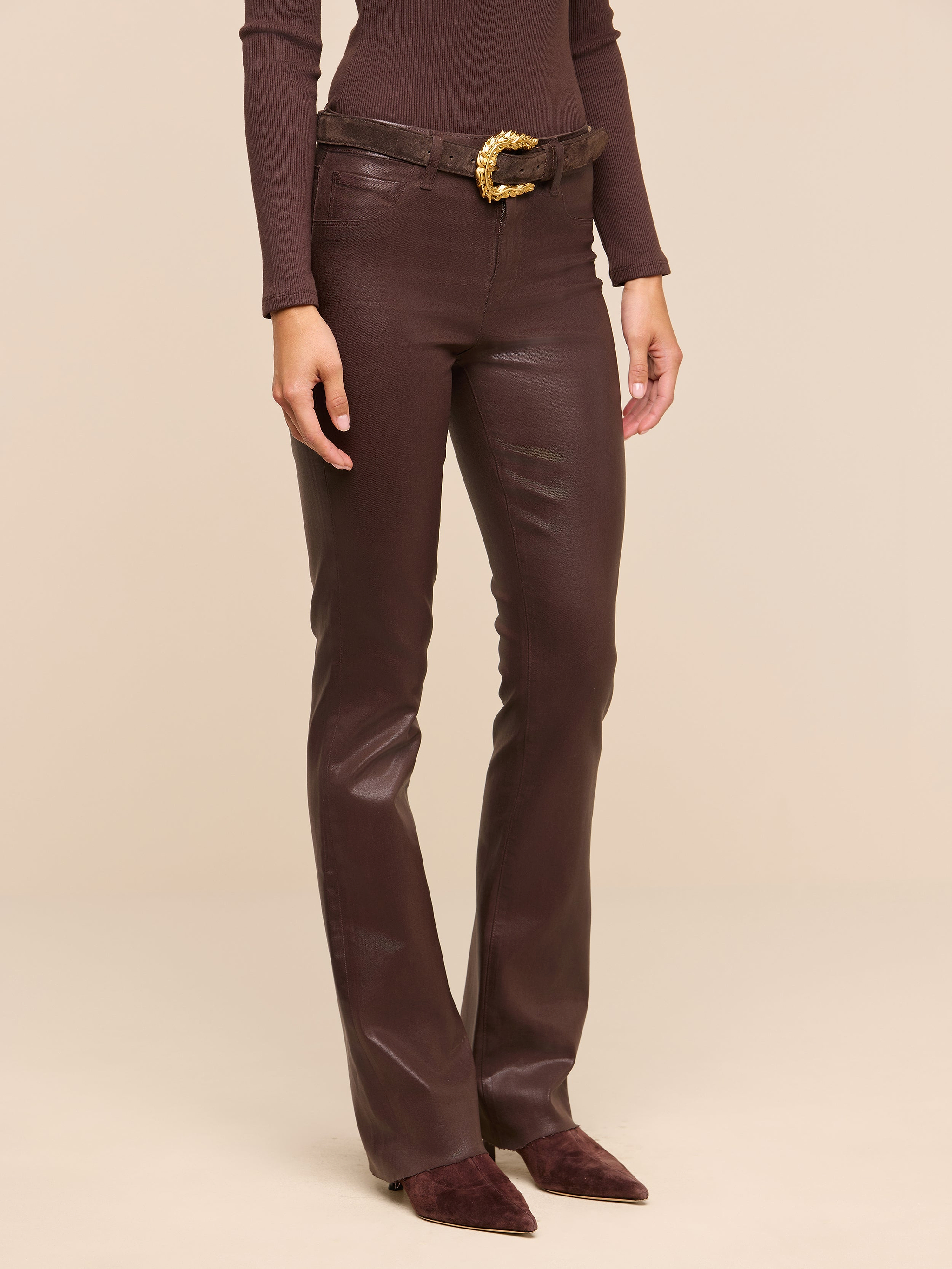 Wearing the Ruth Coated Straight-Leg Jean in brown, along with a coordinating brown long-sleeve top, a brown belt featuring a gold buckle, and pointed brown shoes, a person stands against a plain white background.