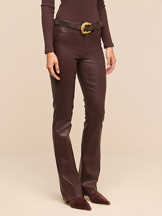 A person wearing the Ruth Coated Straight-Leg Jean in brown, paired with a matching brown long-sleeve top, a brown belt featuring a large gold buckle, and pointed brown shoes, standing against a beige background.