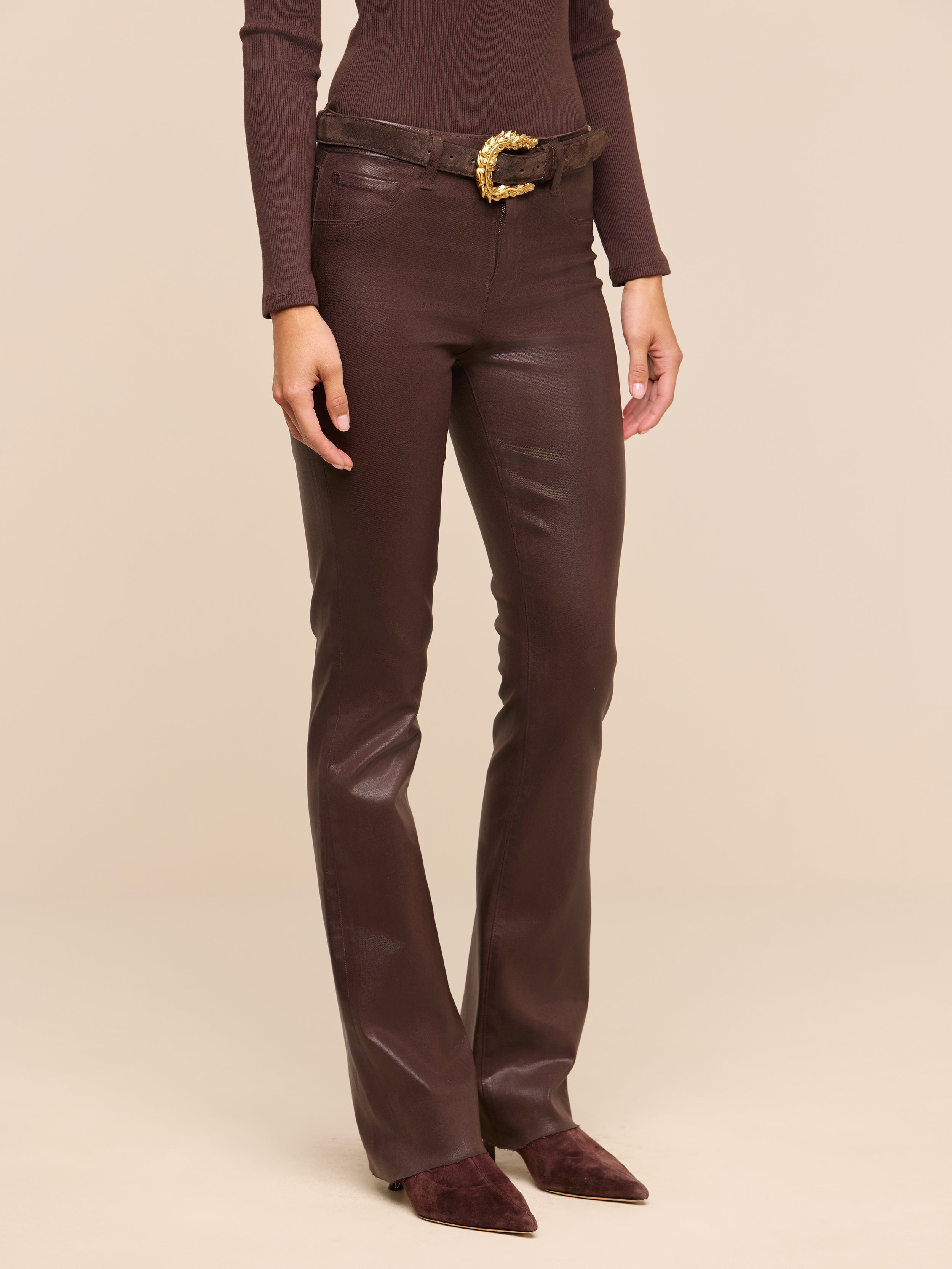 Wearing the Ruth Coated Straight-Leg Jean in brown, along with a coordinating brown long-sleeve top, a brown belt featuring a gold buckle, and pointed brown shoes, a person stands against a plain white background.
