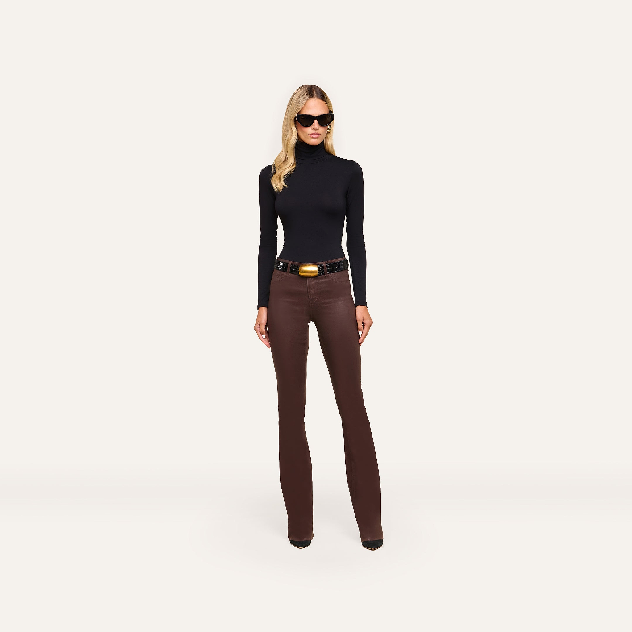 A person wearing a black turtleneck, a black belt with a large gold buckle, and Ruth Coated Straight-Leg Jean high-rise, straight-leg coated stretch denim jeans.