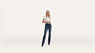 Woman standing against a plain white background, wearing a white t-shirt, dark blue jeans, and black shoes, looking forward with her hands loosely clasped in front of her.
