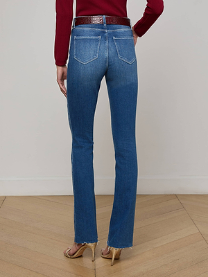 Someone wearing L'AGENCE Ruth Straight-Leg Jean with frayed hems, back pockets, a blue high rise fit, a red long-sleeve top, and heeled sandals stands on a wooden floor facing away from the camera.